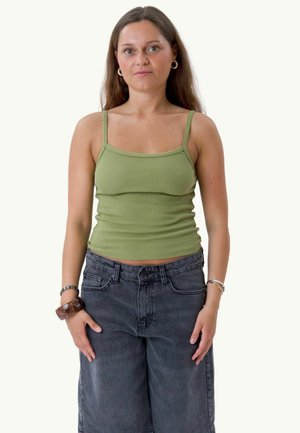 Femme aux longs cheveux bruns portant un débardeur vert, un jean taille haute gris foncé à jambes larges et des bracelets aux deux poignets.
