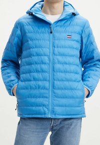 Doudoune bleue avec capuche, présentant un design matelassé horizontal, fermeture éclair à l'avant et logo de la marque sur la poitrine. Matière douce et légère.