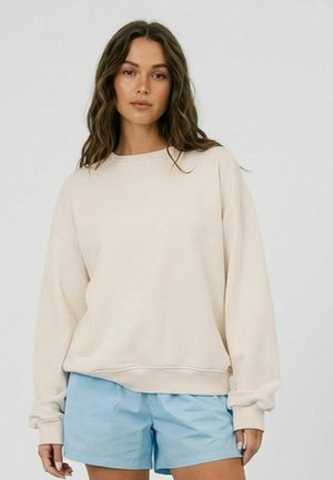 Kobieta o długich falowanych włosach, ubrana w luźną, kremową bluzę i jasnoniebieskie, casualowe szorty, na tle prostego jasnego tła.