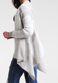 Lehký pletený cardigan v bílé a béžové barvě s diagonálními pruhy, asymetrickým lemem a dlouhými rukávy. Kapsy nejsou viditelné.