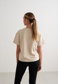 Beige Baumwoll-T-Shirt mit kurzen Ärmeln, lockerer Passform, runder Ausschnitt, getragen mit schwarzen gerippten Leggings. Der Hintergrund ist eine minimalistische weiße Wand.