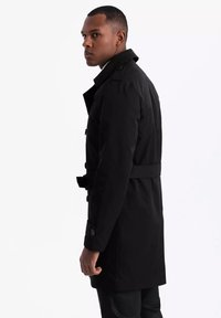 Mężczyzna stojący bokiem, ubrany w czarny płaszcz trench z zapiętymi mankietami i paskiem, na gładkim białym tle.