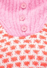 Ružový pletený odev má rebrovanú textúru, tri ružové gombíky a dizajn oranžových hviezd na bielom pozadí.