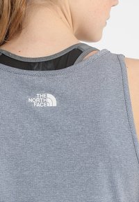 Šedý, strukturovaný sportovní tank top s černou síťovinou pod ním. Obsahuje malé bílé logo na horní části zad. Bez rukávů.