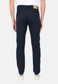 Pantaloni in cotone blu navy con vestibilità slim, dotati di due tasche posteriori e una leggera toppa in pelle beige all'elastico in vita. Texture liscia, design minimal.