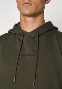 Sweatshirt verde-oliva feito de tecido macio. Apresenta um capuz com cordão e o logótipo bordado "BJÖRN BORG" em azul-marinho. Design simples com detalhes em malha rib o.