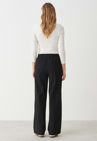 Zwarte wijde broek met een elastische tailleband, voorzien van een gladde textuur en een minimalistisch ontwerp. Witte lange mouwen top is gedeeltelijk zichtbaar.