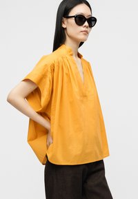 Femme portant un chemisier ample jaune moutarde à manches courtes et des lunettes de soleil noires, debout avec une main dans la poche devant un fond uni.