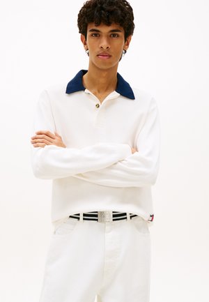 Giovane uomo con capelli ricci che indossa una camicia bianca a maniche lunghe con colletto blu navy, pantaloni bianchi e una cintura a righe bianca e nera, con le braccia incrociate.