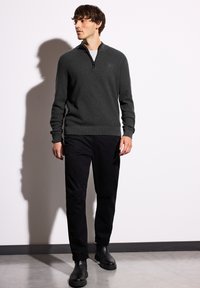 Un pull en tricot gris foncé avec un col zippé à quart, associé à un pantalon noir et des bottines noires. Texture lisse, design ajusté.