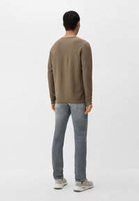 Camicia marrone a maniche lunghe con una finitura texture, abbinata a jeans azzurri e sneakers grigie, vista da dietro su uno sfondo bianco.