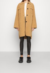 Cappotto oversize sabbia e testurizzato con grandi bottoni e tasche laterali, abbinato a leggings in pelle nera e scarpe slip-on nere.