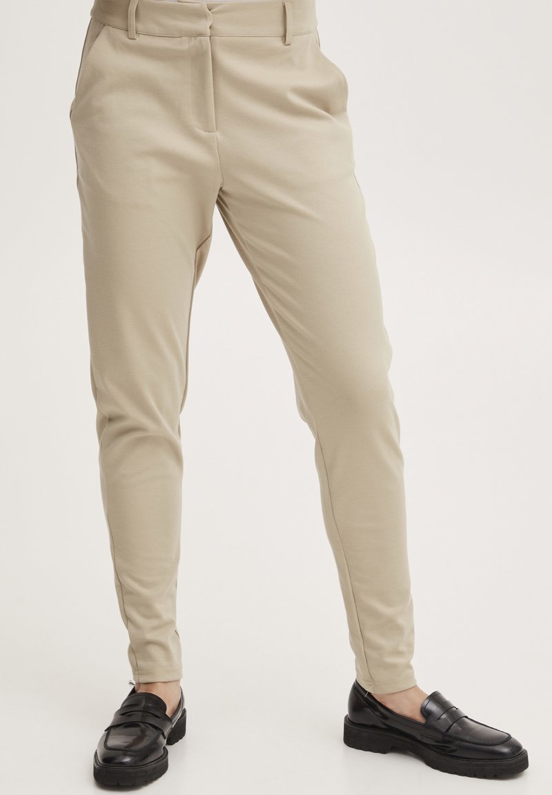 Beige housut, joissa on kapea istuvuus, hoikka muoto, sileä pinta ja etupinnat. Yhdistetty mustiin loafer-kenkiin, joissa on kiiltävä pinta.