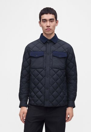 Jeune homme portant une veste matelassée bleu foncé avec des poches poitrine boutonnées et un col en velours côtelé, debout sur un fond blanc uni.
