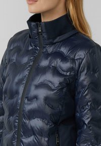 Femme portant une veste matelassée bleu marine brillante avec un col haut côtelé et une fermeture éclair frontale visible, montrée de la nuque à la taille.