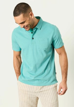 Mężczyzna w zielonkawo-niebieskiej koszulce polo z krótkim rękawem i kieszenią oraz beżowych spodniach w paski, patrzy w dół i uśmiecha się z rękami w kieszeniach.