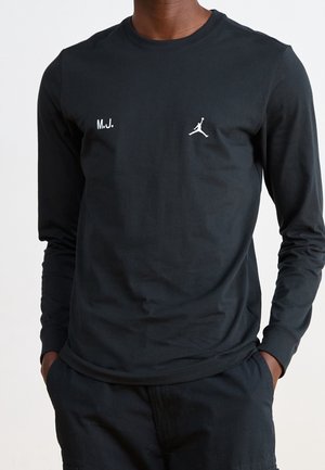 Mężczyzna ubrany w czarną koszulkę z długim rękawem z białym napisem "M.J." i logo Jumpman na piersi, ręce w kieszeniach czarnych spodni.