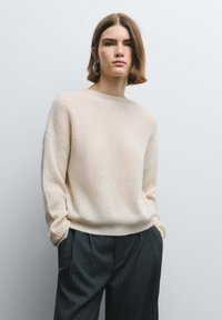 Maglione beige lavorato a maglia con scollo rotondo, maniche lunghe larghe e trama a coste, abbinato a pantaloni scuri a righe sottili.
