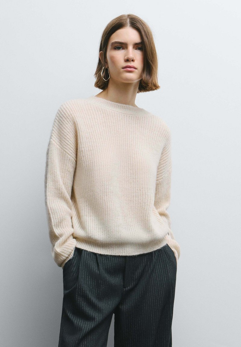 Maglione beige lavorato a maglia con scollo rotondo, maniche lunghe larghe e trama a coste, abbinato a pantaloni scuri a righe sottili.