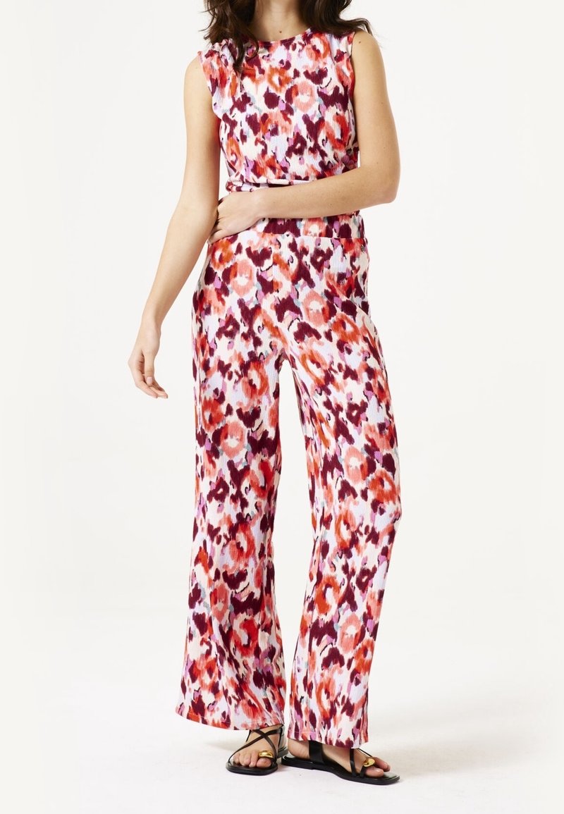 Kombinezon w kwiatowy wzór w odcieniach różu, pomarańczy i fioletu, z dopasowaną górą, szerokimi spodniami i teksturowaną tkaniną. Płaskie czarne sandały.