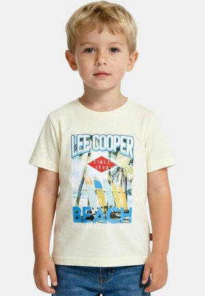 Garçon blond portant un T-shirt crème avec l'inscription "Lee Cooper Beach" et un motif de surf, palmier, associé à un jean bleu.