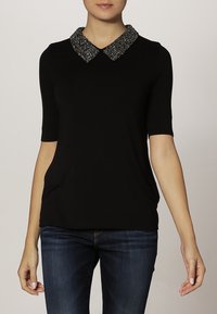 Kvinna i svart kortärmad topp med en texturerad glittrig krage, kombinerad med mörkblå jeans, stående mot en neutral bakgrund.