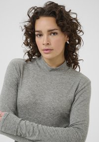 Gray ribbed long-sleeve top with a mock neck. Soft fabric, fitted shape, and minimal stitching details. Model has curly hair and is posed with arms crossed.