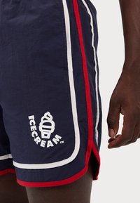 Marineblå shorts med røde og hvide sidelister, der har et isvaffel-logo. Stoffet synes let og har en glat tekstur.