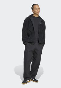 Chaqueta de forro polar negra con cremallera y capucha, combinada con pantalones cargo negros. Los detalles notables incluyen bolsillos laterales y franjas blancas en contraste en los zapatos.