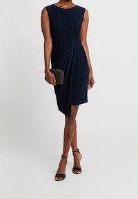 Marinblå ärmlös klänning med draperad textur, knälång klippning och sidoknytdetalj, i kombination med en svart clutch och svarta sandaler med remmar.