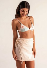 Top de bikini floral en azul claro, con acentos melocotón; combinado con una falda envolvente arrugada en color crema, atada en la cintura con flecos. Fondo plano.