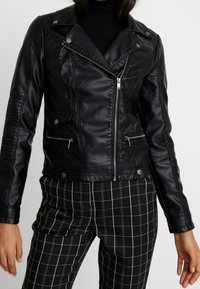 Sort motorcykeljakke i imiteret læder med lynlåsede lommer, skulderstropper og quiltede skulderdetaljer, kombineret med sort-hvide ternede bukser.