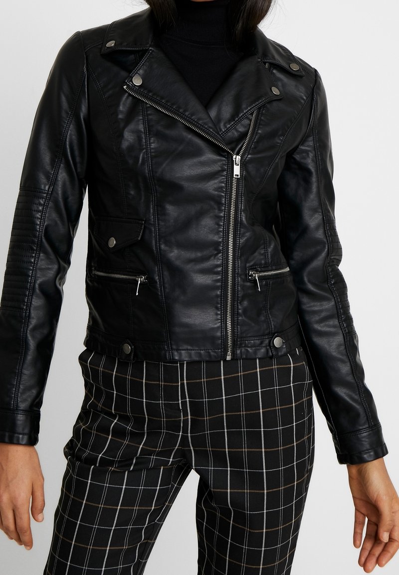 Czarna motocyklowa kurtka z ekoskóry z zamkami błyskawicznymi na kieszeniach, pagonami i pikowanymi detalami na ramionach, zestawiona z czarno-białymi szachownicowymi spodniami.