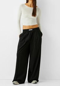 Mustat, leveät housut, joissa on kiristysnauha vyötäröllä, yhdistettyinä tiukkaan, vaaleanharmaaseen pitkähiharaiseen paitaan. Sisältää sivutaskut ja merkkikorosteen.