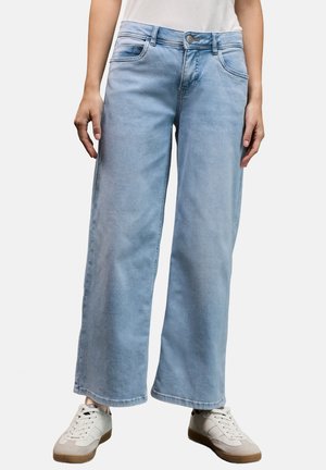 Jean large bleu clair en denim avec une fermeture à bouton, cinq poches et un léger effet dégradé, associé à des baskets blanches.