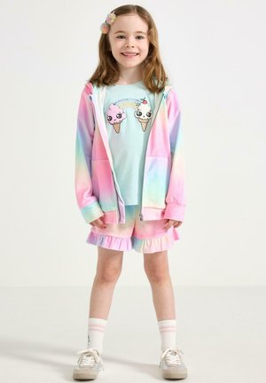 Unge pige smiler, iført pastel regnbue hættetrøje, matchende flæse-shorts, lyseblå skjorte med tegneserie isvafler, hvide sokker og sneakers.