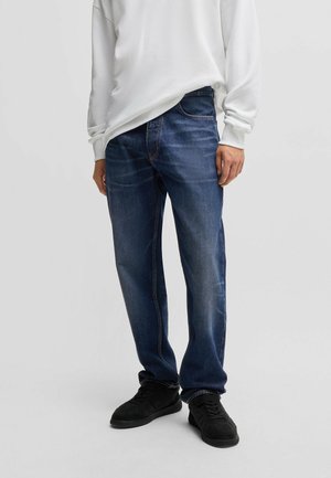 Dark blue denim jeans with a straight leg design, five pockets, and contrast stitching, paired with a white long-sleeve top and black sneakers.