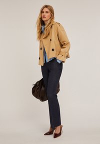 Beige dubbelgebreide trenchcoat met zwarte knopen, lichtblauw shirt, donkere broek en bordeauxrode puntige hakken, aangevuld met een bruine tas.