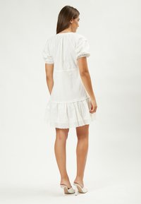 Weißes Baumwollkleid mit kurzen Puffärmeln, gestuftem Rüschensaum und Spitzenverzierungen am Saum, kombiniert mit weißen offenen Absatzschuhen.