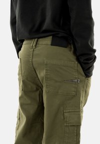 Pantaloni cargo verde oliva con multiple tasche, una finitura testurizzata e un'etichetta nera in vita, abbinati a una maglietta nera a maniche lunghe.