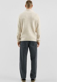 Beige geribbelde trui met een kraag, gecombineerd met donkergrijze, losvallende jeans. Voorzien van subtiele stiksels en schuine achterzakken.