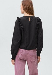 Blusa nera con spalline arricciate e tessuto texturizzato, abbinata a jeans rosa con dettagli a forma di stella sulle tasche posteriori.