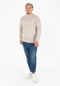 Felpa beige con logo "Timberland" in bianco, abbinata a jeans blu e sneakers bianche. Tessuto liscio, collo a giro, maniche lunghe.