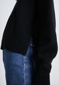 Maglione lavorato a maglia nero con polsini e orlo a coste, abbinato a jeans blu. Presenta una vestibilità comoda e una texture morbida, visibile in vista laterale.