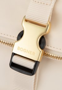 Beige taske med spænde i guld og sorte detaljer. Har en glat tekstur og en lynlåslukning langs syningen. Logo synligt på spændet.