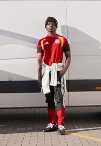 Athletic outfit: red shirt with yellow accents, black sleeves, black and red pants, white sweater tied around waist, black beanie, black shoes.