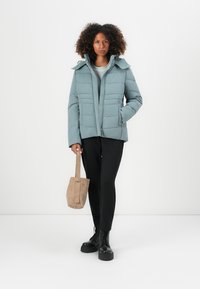 Lyseblå pufferjakke med høj krave, med lynlås og trykknapslukninger, båret over en grå sweater og sorte bukser, med en beige taske.
