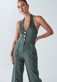 Olivově zelený overal s hlubokým výstřihem do V, předními knoflíky, střih na míru a širokými nohavicemi s bočními kapsami.