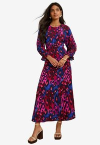 Maxi-Kleid mit einem kräftigen Leopardenmuster in Pink, Lila und Blau. Hat lange, ausgestellte Ärmel und eine taillierte Taille. Getragen mit Sandalen mit Blockabsatz.
