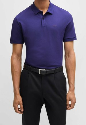 Mand iført en tætsiddende mørkelilla polo shirt med "BOSS"-logo og sorte bukser med sort bælte, stående mod en ensfarvet baggrund.